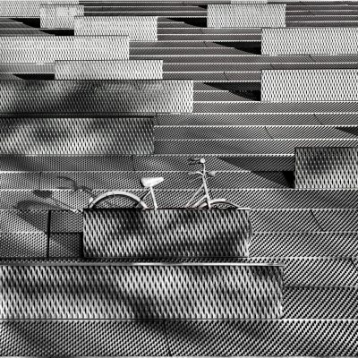 A black and white photograph of an urban bike park made from a grid pattern metal mesh with a geometric design. A bicycle is parked on one level, surrounded by patterns of straight lines and angles. The background features multiple levels with different sizes and shapes of rectangular cells, creating intricate shadows that enhance the modern aesthetic. This scene combines industrial elements like steel structures with artistic details to create a visually striking composition in the style of a modern geometric design.