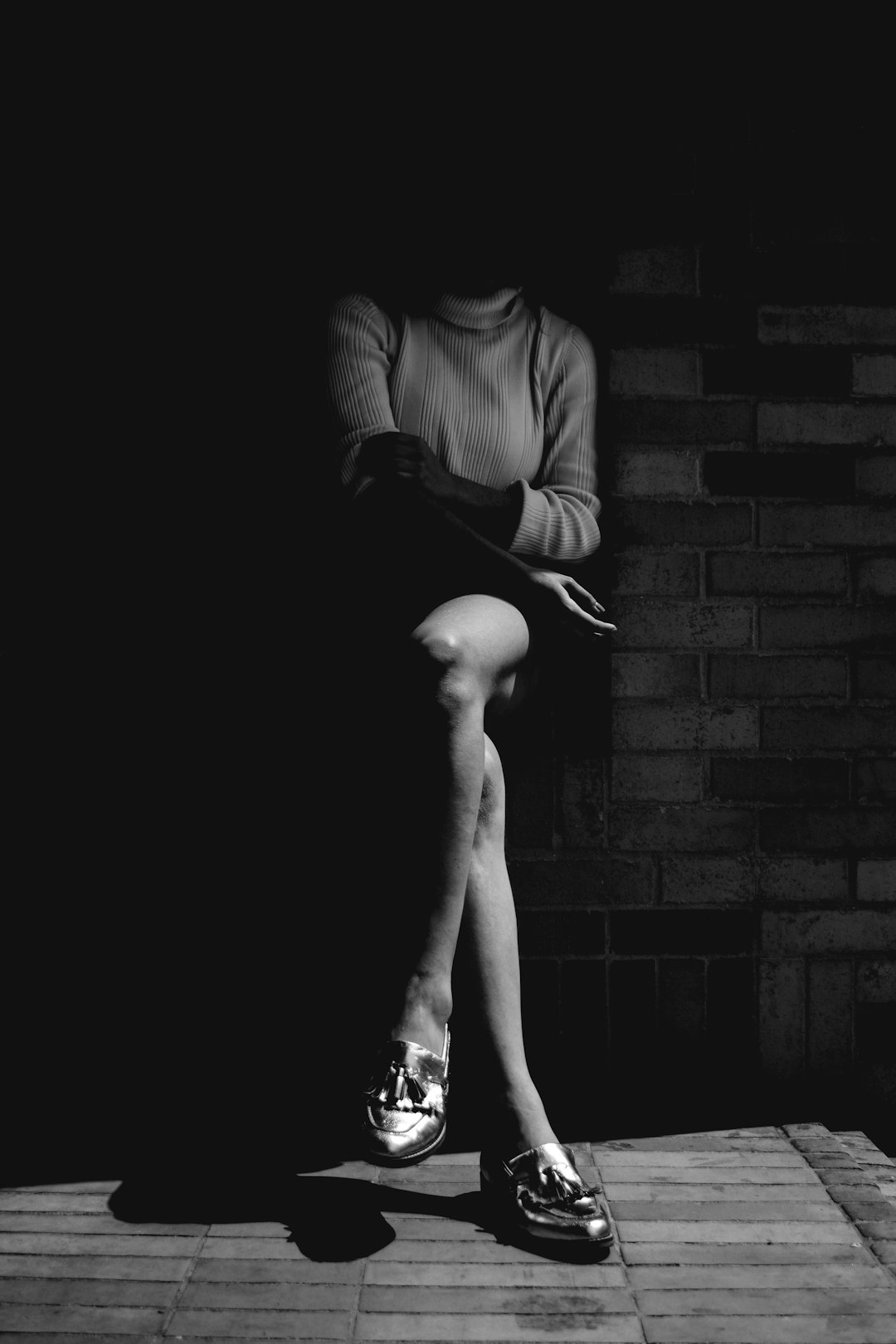 A black and white portrait of an attractive woman sitting on the edge of her shoes, legs crossed, wearing a sweater and skirt, golden loafer shoes, against a dark brick wall. Soft lighting creates deep shadows in front of her face. Shot with Leica M6 using Kodak TriX film. The atmosphere is mysterious. High contrast, lowkey photography style in the style of emphasizing textures and shapes in chiaroscuro lighting. –ar 85:128