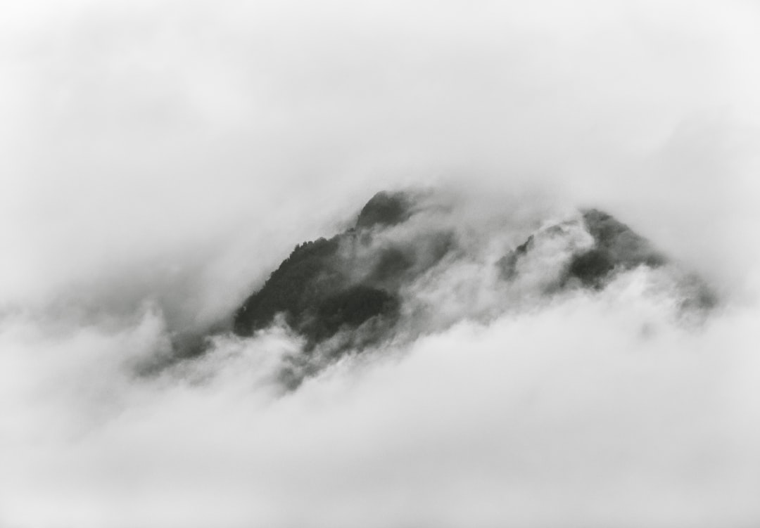 Black and white photography of a mountain peak emerging from clouds in a minimalist, high contrast, low angle shot. The monochrome photo has soft lighting and a foggy atmosphere. The nature-inspired artwork has a simple yet powerful composition featuring natural elements with a misty effect and subtle textures on the cloud formations. –ar 128:89