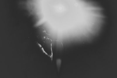 A blurry black and white photo captures a moment in music photography style. It shows the silhouette of someone singing into a microphone on stage, illuminated by a spotlight. The background is dark with subtle hints of lights or silhouettes, creating depth and contrast. This photograph conveys raw emotion as they sing along to one song after another at a live concert in the style of raw emotion. --ar 128:85