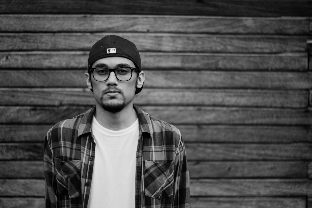 Black and white portrait of a young man with glasses, wearing a baseball cap and flannel shirt over a t-shirt, standing against a wooden wall. Shot in the style of Sony A7R III raw. –ar 128:85