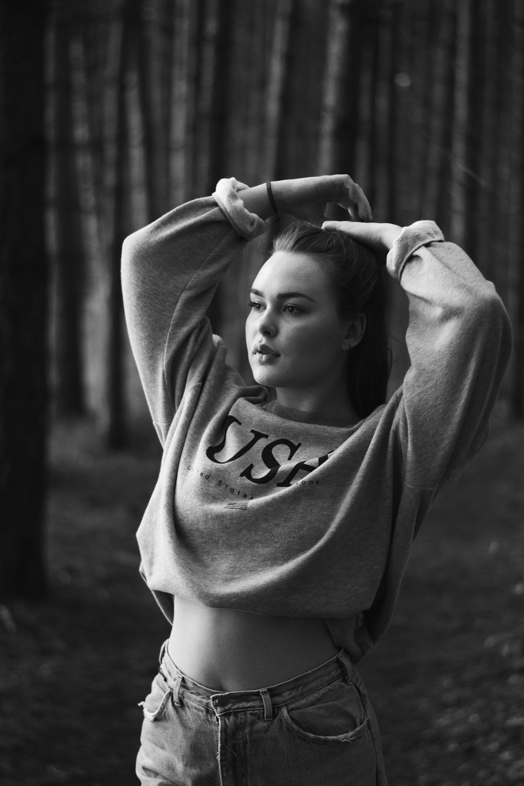 A gorgeous woman with her hands behind her head wearing an oversized sweatshirt that says “usm” on the front, standing in a forest in black and white photography with high contrast, shot on a Fujifilm Superia in the style of a Hasselblad X2D. –ar 85:128