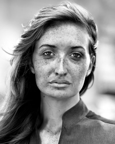 black and white portrait of an elegant woman with freckles, facing the camera directly, in Paris street, wearing modern business attire, long hair, shot on Leica M6 using natural light to capture her features and personality, her expression is one that reflects confidence and determination, the background is blurred to focus attention on subject, the overall mood conveys elegance and strength --ar 51:64