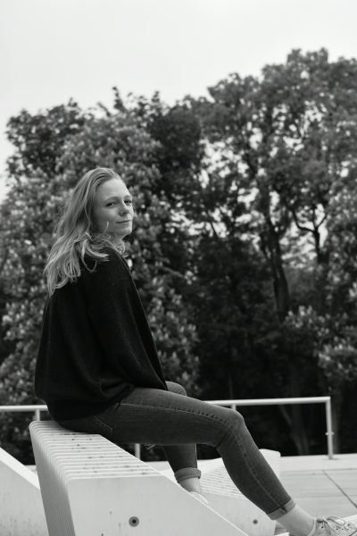 Black and white street photography of a blonde woman sitting on the edge of a concrete platform, wearing a black sweater with jeans, smiling at the camera, with trees in the background, white sneakers, on a summer day, in the style of Leica M6. --ar 85:128