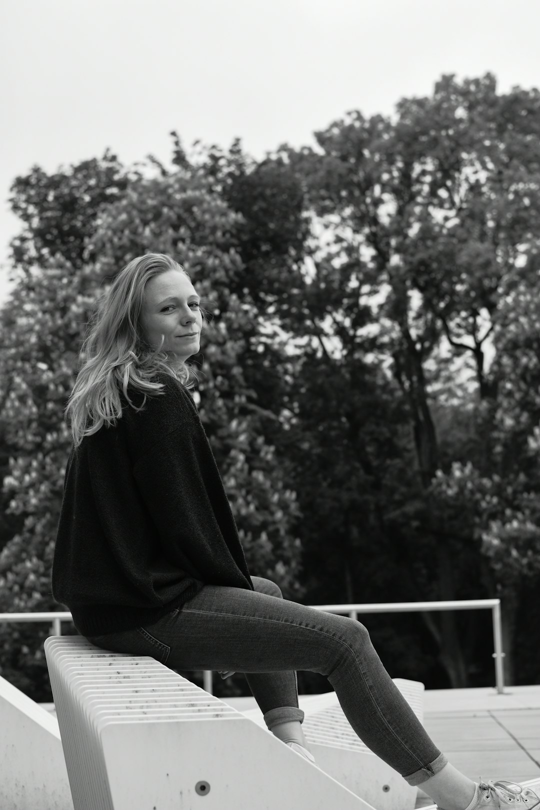 Black and white street photography of a blonde woman sitting on the edge of a concrete platform, wearing a black sweater with jeans, smiling at the camera, with trees in the background, white sneakers, on a summer day, in the style of Leica M6. –ar 85:128