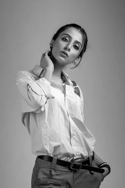 A young woman with her hair tied back, wearing an unbuttoned white shirt and gray trousers with suspenders posing while holding one arm behind her neck and looking at the camera in front of a gray background, monochrome black & white, hyper realistic photography captured in the style of Canon EOS R5 C. --ar 85:128