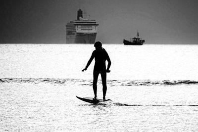 Black and white photo of a surfer standing on a surfboard in the middle of the ocean, with a cruise ship far away in the background as a silhouette. The photo is in the style of a silhouette. --ar 128:85