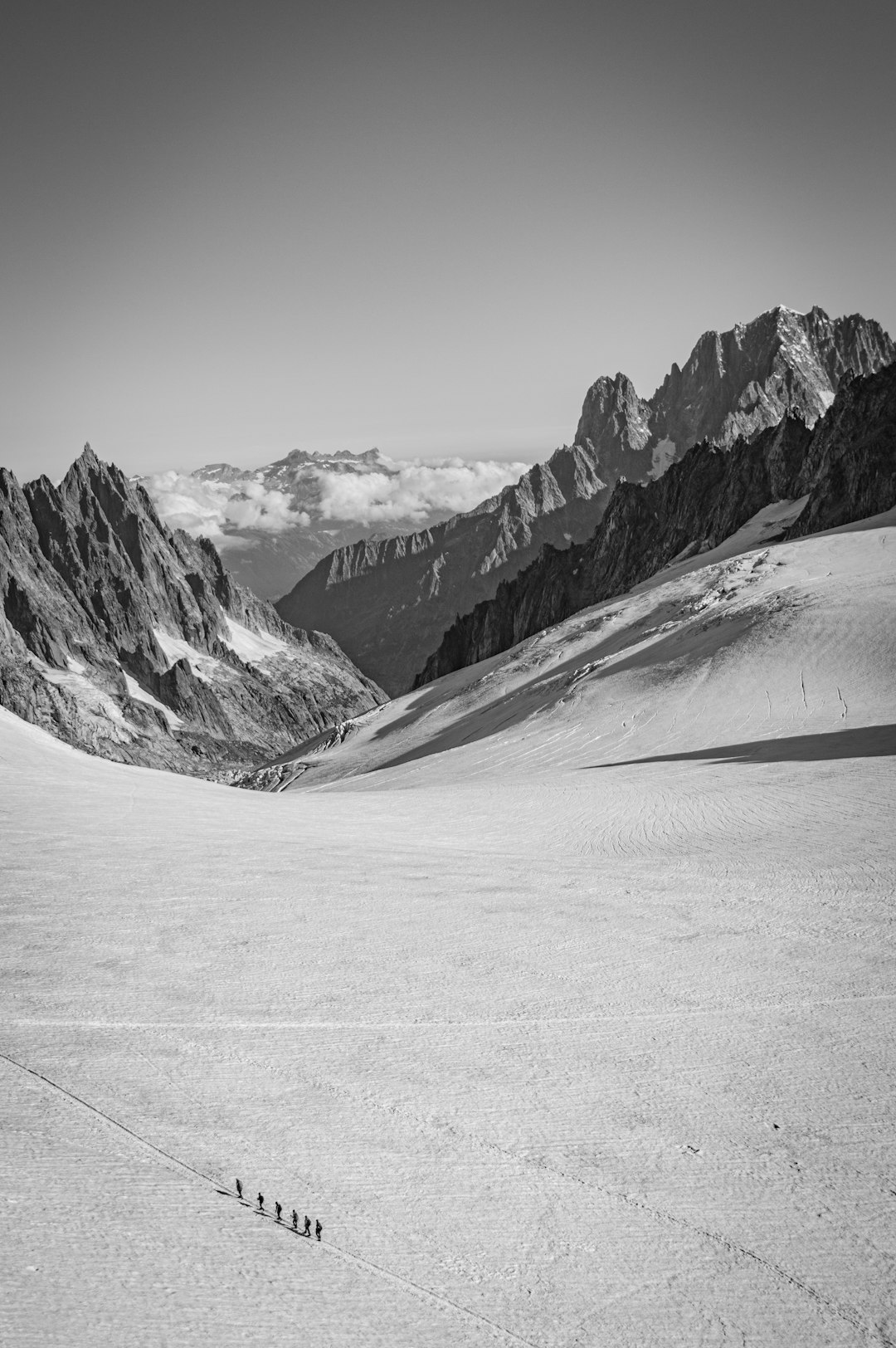 Black and white photography of an alpine snowfield with skiers walking up the side, large mountains in the background, wide shot, Leica M6 camera, 28mm lens, f/4 aperture, natural daylight, cool tones, sharp focus on details, subtle contrast between snowy peaks and clear sky, capturing the sense of adventure in the extreme environment. in the style of natural daylight. –ar 85:128