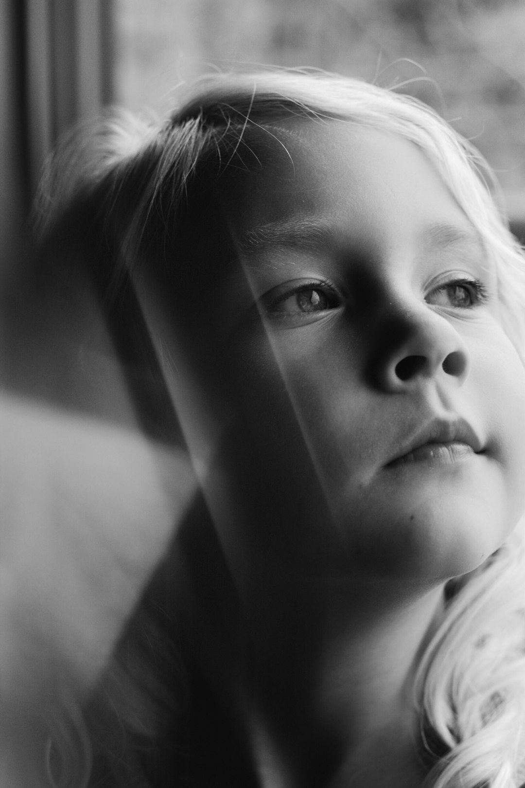 A black and white portrait of an eight-year-old girl with blonde hair, her face partially obscured by the edge of a window. The focus is on capturing her expression as she gazes out through the glass. Soft natural light illuminates her features, creating gentle shadows that add depth to the scene. This photo was taken using a Fujifilm XT4 camera and a Fujinon XF lens, adding warmth and texture to create a timeless moment frozen in time in the style of an early 20th century portrait photographer. –ar 85:128