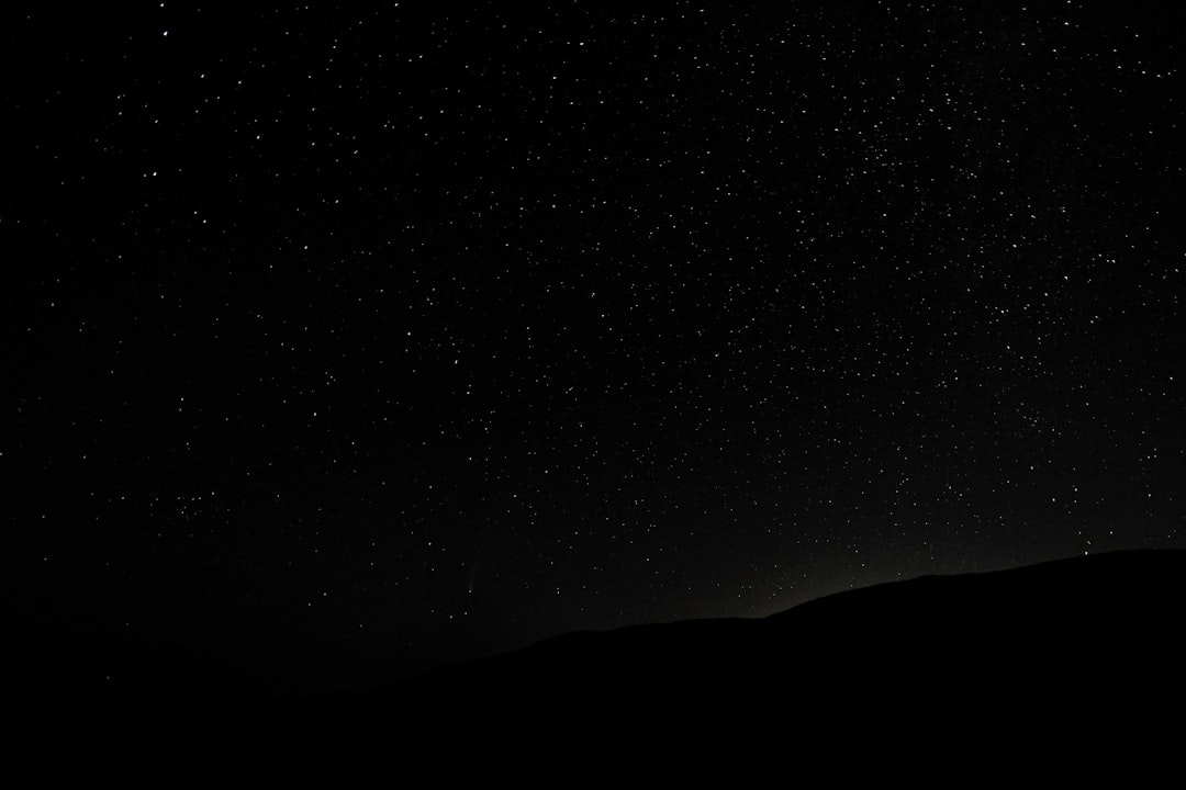 A black sky with stars, at night, a dark silhouette in the top left corner, distant mountains in the bottom right corner, high resolution, high quality, high detail, super realistic, cinematic, professional photography, shot with a canon r3 camera, depth of field, volumetric light, natural light, in the style of no artist. –ar 128:85