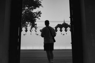 A black and white photo of an athlete in shorts standing at the entrance to Bangkok's royal palace, seen from behind with his back turned, holding onto his phone, looking out into people playing football on the field outside, shot in the style of a Canon EOS R5 F2 ISO40 36mm. --ar 128:85