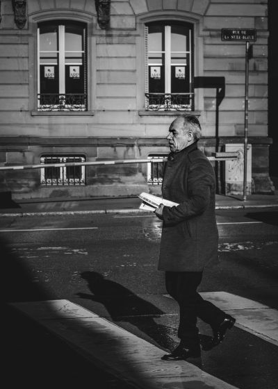 black and white street photography of an elegant middle-aged man walking on the sidewalk in Paris, holding his book under one arm while he reads it with intense focus. High contrast, natural lighting creates shadow play in the style of Leica Q2 Monochrom with a Summilux f/8 lens. --ar 91:128