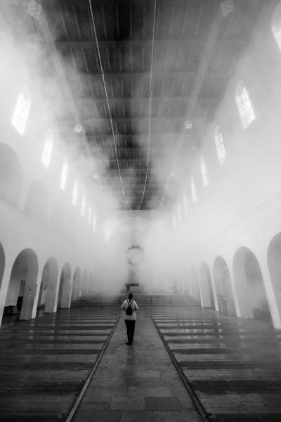 A woman is walking in the church in a foggy atmosphere. It is black and white photography with a symmetrical composition captured with a wideangle lens in soft light creating a mysterious mood. The photo was taken with a Sony Alpha A7 III in the style of black and white photography. --ar 85:128