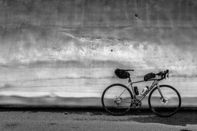 Black and white photography of an unbranded road bike leaning against the wall with a black duffle bag on its back, taken from far away. The bicycle is standing next to it, with lots of space in between them. It's in front of a concrete wall, with a lot of grain. The photo is in the style of an anonymous photographer. --ar 128:85