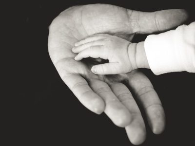 A black and white photograph shows an adult hand holding small fingers, palm up, with a baby's tiny hands resting on it. The plain black background highlights their connection in this tender moment. The high resolution photography captures insanely detailed and fine details in a hyper realistic style. --ar 4:3