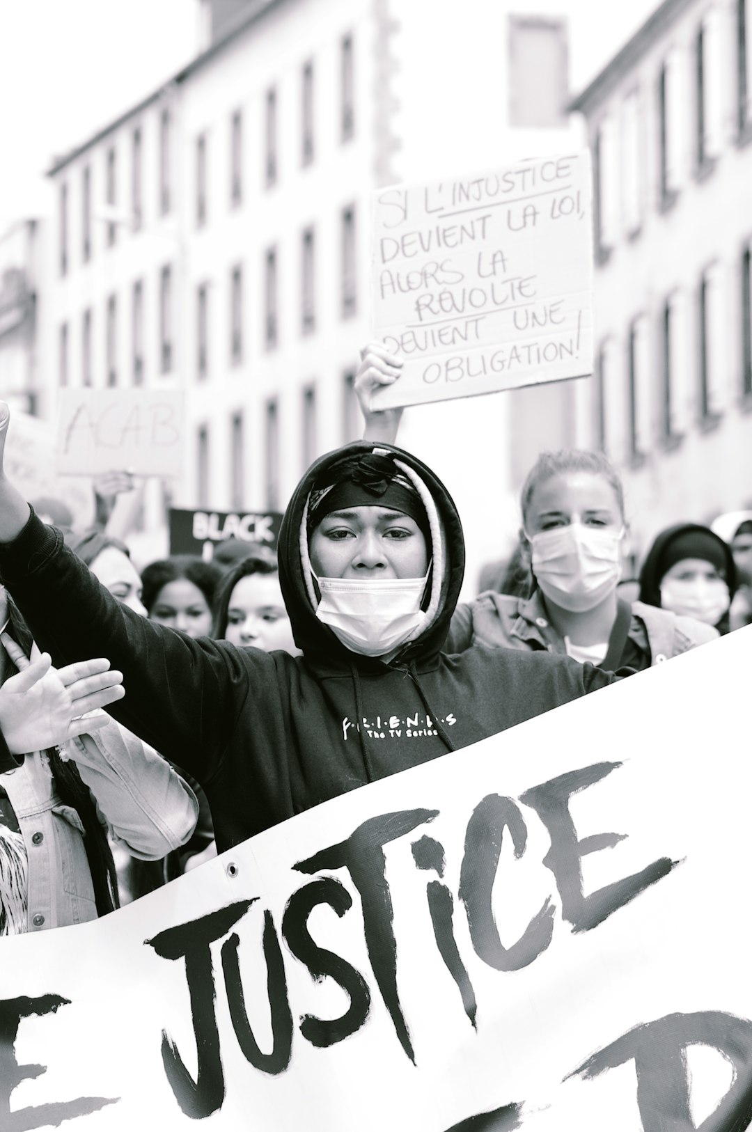 Black and white photography of a French woman wearing a black hoodie with a face mask holding up a banner saying “hidden justice” in front, surrounded by other people on the street, some holding signs reading ‘right to be free’, ‘seaside’, with a French revolution theme, very detailed faces, a low angle shot, taken with a wideangle lens, at an f/4 aperture, using natural daylight, in the style of a documentary photographer. –ar 85:128