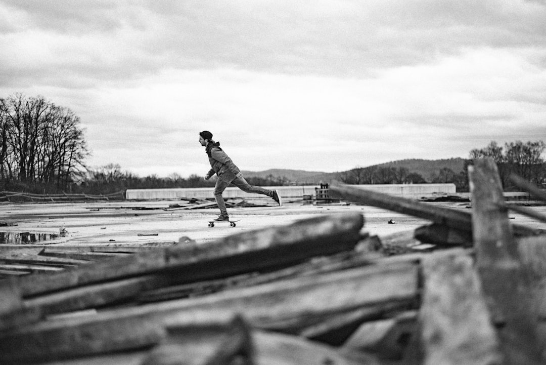 A black and white photo of an ice skater gliding on wood planks in the middle of a construction site, woods behind him, distant hills, overcast sky, medium shot, 35mm film grain, f/24, Kodak TMax film. –ar 128:85