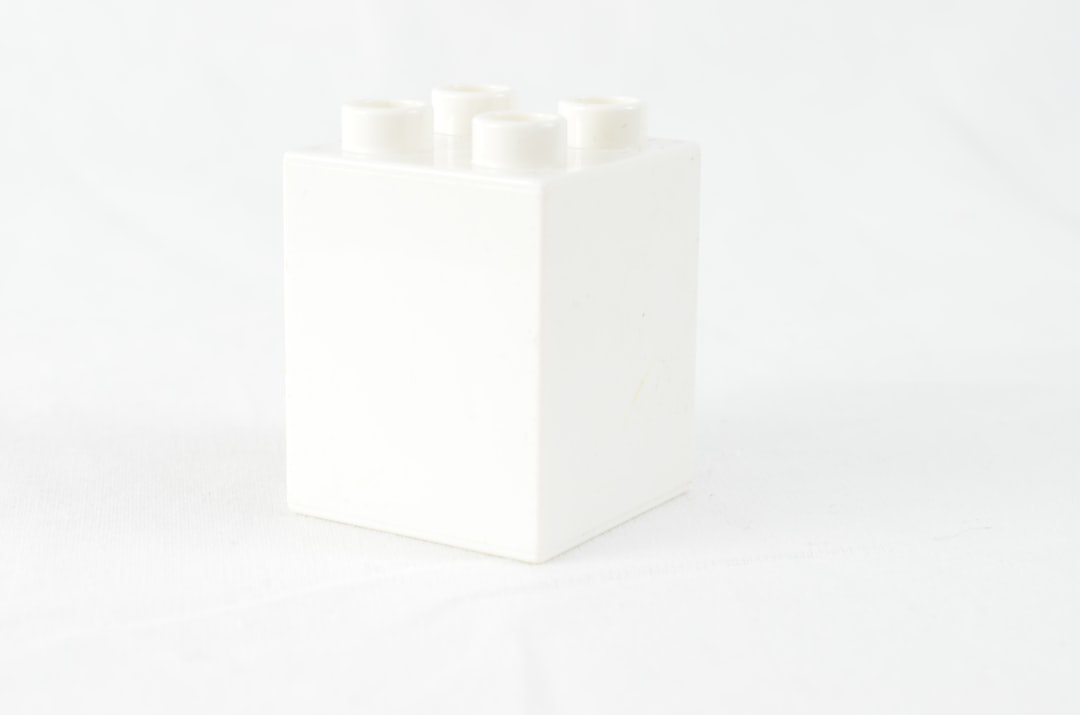 A white LEGO brick sits in the bottom left corner of an all-white background. The square-shaped brick has four rounded edges on each side. It is made of matte plastic material with a smooth surface and features a solid color that reflects the light in its surroundings. This scene highlights minimalism in the style of LEGO bricks. –ar 32:21