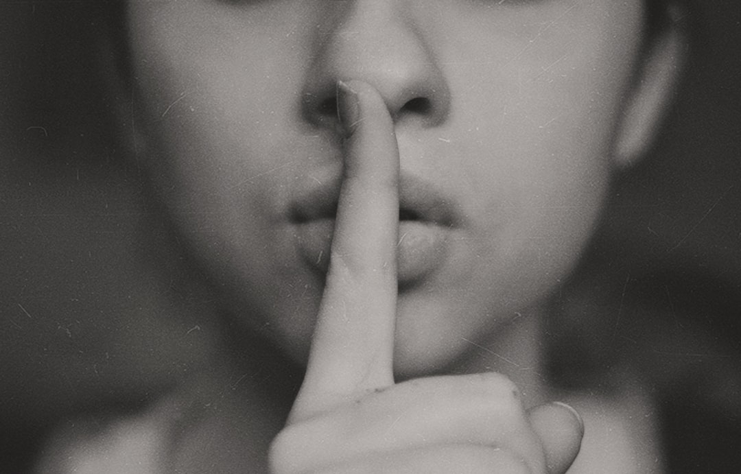 A person with their index finger placed to the corner of their mouth, creating a [HUSH](https://goo.gl/search?artist%20HUSH) gesture, indicating silence or navigating within another’s space. The focus is on the face against a monochrome background in the style of a vintage style with a grainy texture and blurred background. It is a black and white closeup shot from a low angle perspective with soft lighting, capturing the nostalgic feel of a vintage film camera and its grainy effect. –ar 128:81