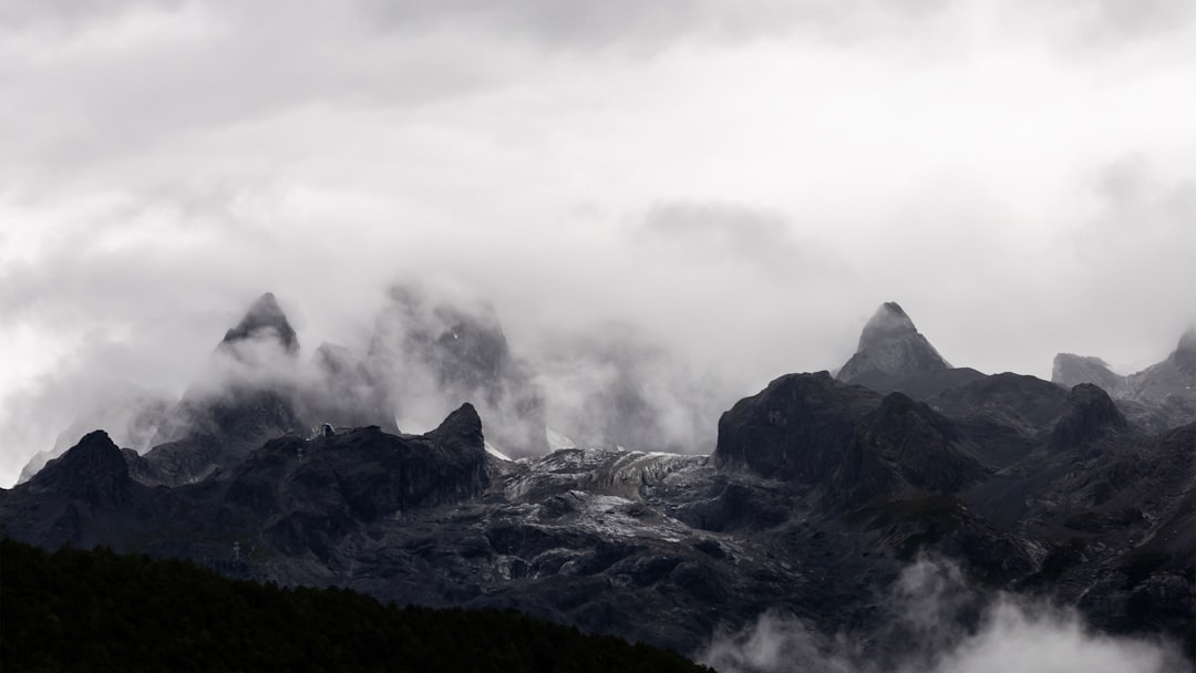 The rugged peaks of the Alps stand tall against an overcast sky, their silhouettes obscured by thick clouds and mist that swirl around them. The scene captures the raw beauty of nature’s grandeur with its towering rock formations, dramatic mountain range, and dense fog creating a sense of mystery and awe in each frame. –ar 16:9