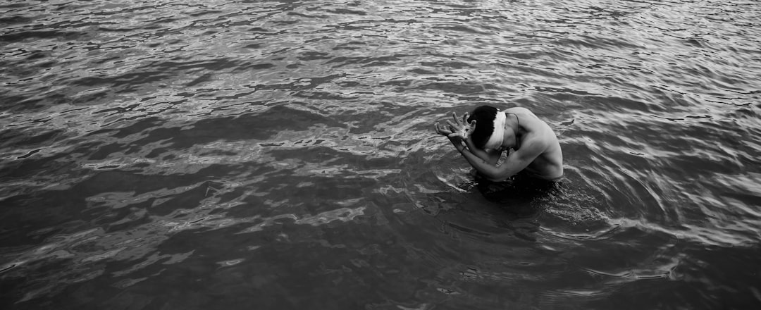 A man is kneeling in the water, holding his head and crying with tears on it. The background of calm sea surface has black and white tones. Shot from above, full body shot, documentary photography, editorial photography, wideangle lens, fstop f/4, ISO200, –ar 32:13