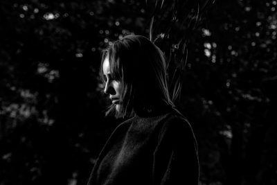 A woman in her thirties, standing alone at night with the silhouette of trees behind her. She is wearing an oversized black sweater and has shoulderlength hair. The lighting casts long shadows on her face as she gazes into distance, adding to his mysterious aura. Black & white photography, using Fujifilm XT4 for a highresolution shot. --ar 128:85