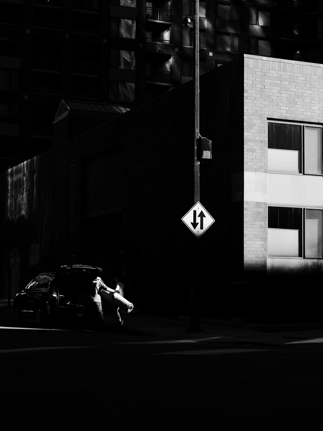 Black and white street photography, minimalism, graphic design, city landscape, shadows, reflections, night time, a woman running in the foreground with an arrow sign pointing to her right on a pole next to a building wall, a parked car, monochromatic. –ar 3:4