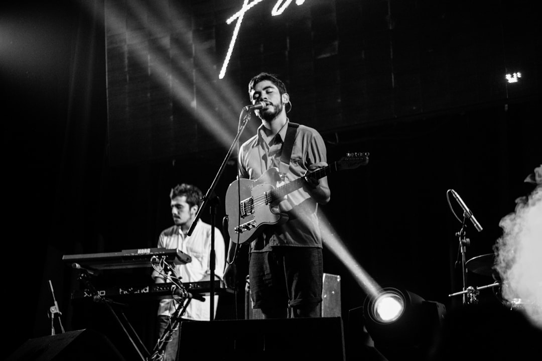 A black and white photograph shows two young men playing music on stage, one man is singing into the microphone while another plays an electric guitar. There is smoke coming from behind them, they stand in front of large speakers that glow with light. They look focused but happy as if it was their dream to play live at such a venue. Shot in the style of [Martin Parr](https://goo.gl/search?artist%20Martin%20Parr) using a Leica M6 camera, the background has dark tones with hints of yellow color. The atmosphere feels excited yet calm. –ar 128:85