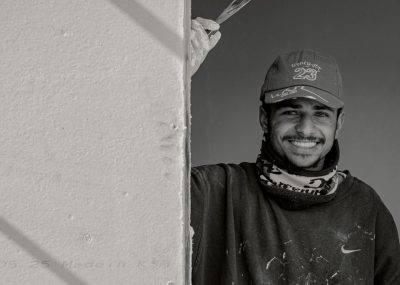 A black and white photo of a smiling young Moroccan man painting on the side wall, wearing a Nike cap and sweatshirt with "925" on it, with a scarf around his neck. The photo was taken with a Canon EF 30mm f/8 lens. The style of painting is in the style of street art. --ar 128:91