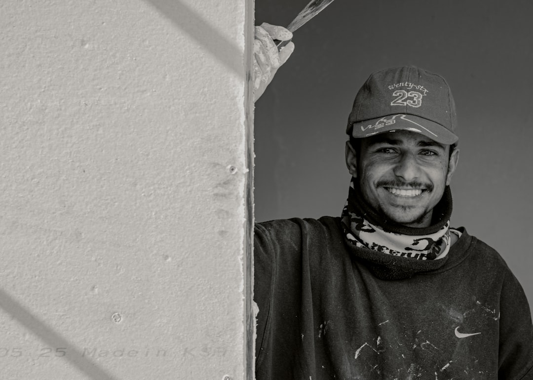 A black and white photo of a smiling young Moroccan man painting on the side wall, wearing a Nike cap and sweatshirt with “925” on it, with a scarf around his neck. The photo was taken with a Canon EF 30mm f/8 lens. The style of painting is in the style of street art. –ar 128:91