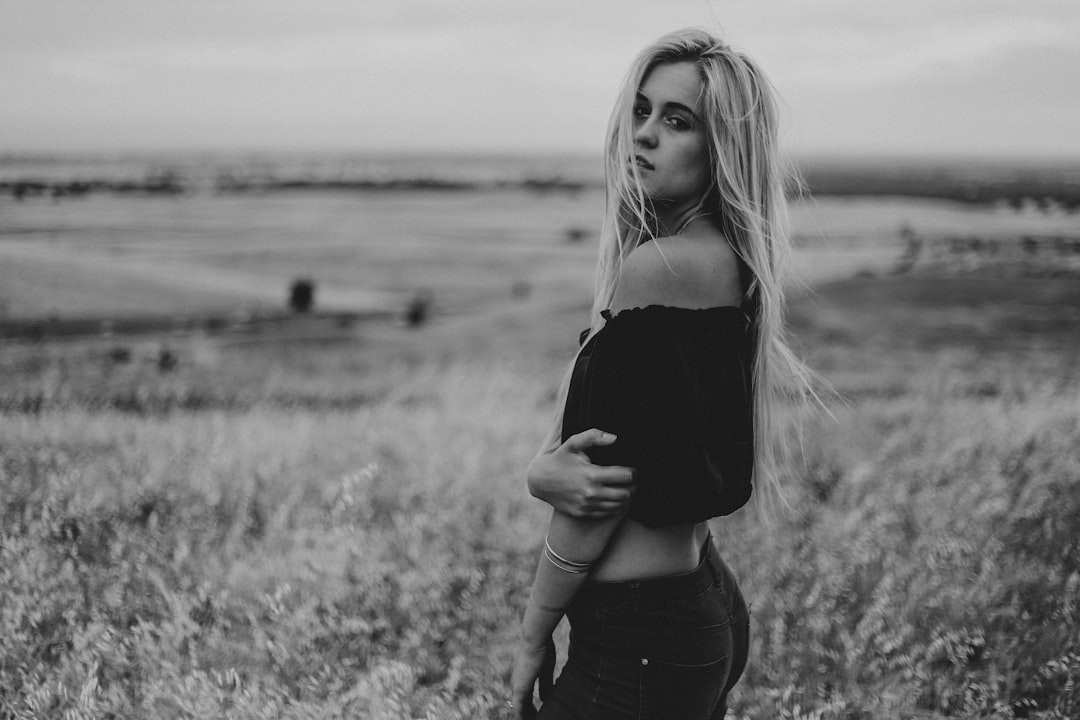 black and white photo of blonde woman posing in the middle of an open field, she is wearing black off shoulder top with jeans, she has her back to camera, she looks over left side, shot on Leica M6 –ar 128:85