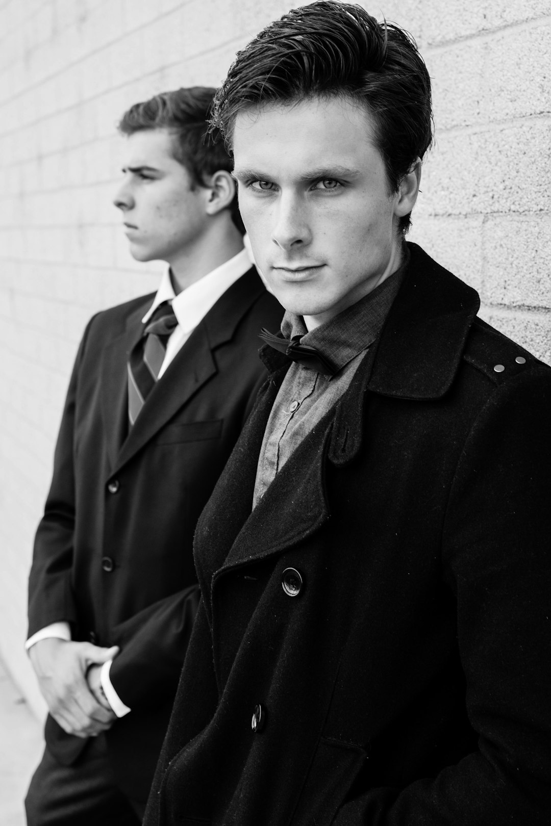 A photo of two men in suits and coats, one with short hair leaning against the wall, the other standing behind him. The man on his right is wearing an elegant coat over a white shirt and tie, while he stands next to another well-groomed young male model. They both have dark eyes and sharp facial features, captured in the style of Canon EOS5D Mark III camera with a black & white filter. –ar 85:128