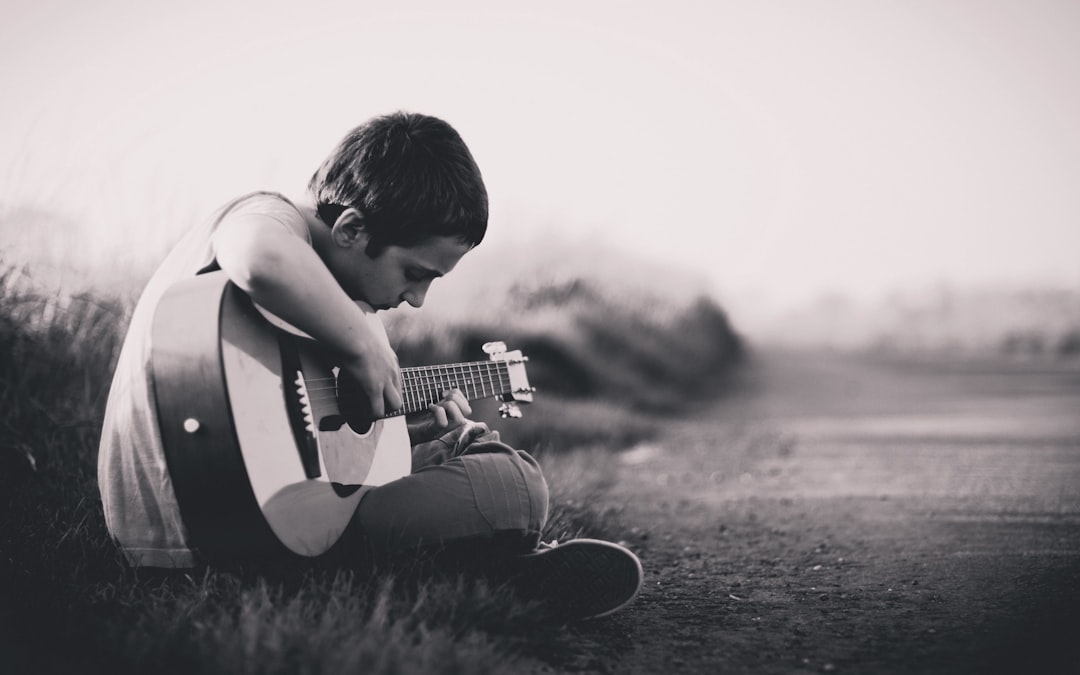 A young boy playing guitar on the side of an empty road, monochrome, high contrast, grainy texture, vintage feel, nostalgic mood, wideangle lens, soft natural light, sitting posture, sad expression. The scene has a vintage feel and nostalgic mood with a monochrome, high contrast and grainy texture captured with a wideangle lens in soft natural light. The young boy sits with a sad expression as he plays guitar on the empty road. –ar 8:5