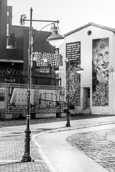 Black and white street photography of an urban art installation featuring graffiti on the side wall, street lamps, corner with cobblestones in front, large murals depicting famous figures such as Steve Jobs, J Robert McCordtson, Elon Musk and Bill Gates, and "A afro poolside" written on one banner. The scene is set against the backdrop of buildings, creating a contrast between old architecture and modern artistic expression. --ar 85:128