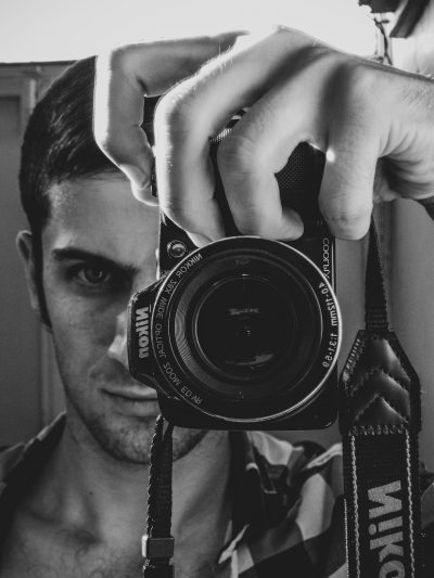 A young man taking a selfie with his Nikon D5 camera, holding the lens in front of him and showing it to us. His face is visible, he has short hair and an athletic build. The photography is in the style of black and white photorealistic hyperbole. --ar 3:4