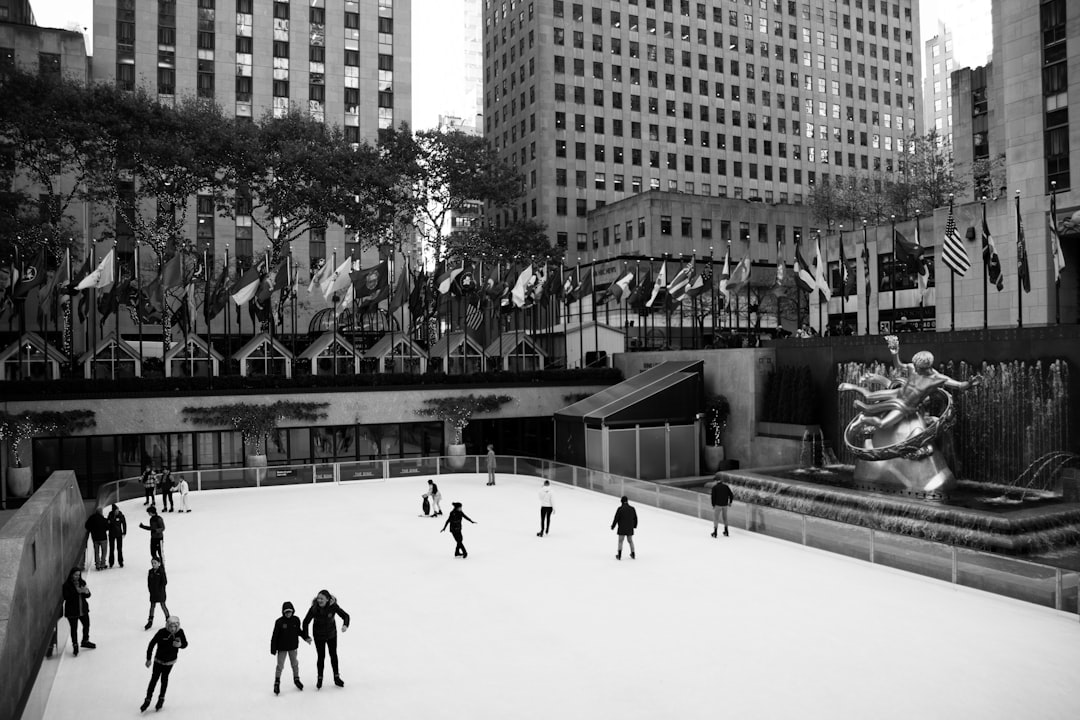 Black and white photo of the ice rink at Rockafeller center in New York, people skating on it, a statue is visible in front of them, there’s also some buildings behind the scene, shot with Sony Alpha A7 III camera, –ar 128:85