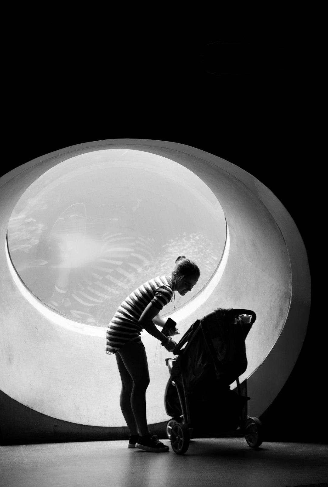 A black and white photograph of an adult woman with short hair in a striped t-shirt, standing next to a baby stroller while putting away a toy inside it, inside the large round entrance of a tunnel tube, with high contrast, soft light, natural lighting, in the style of minimalist style, with a wide-angle lens, sharp details, smooth movements, and warm tones. –ar 43:64