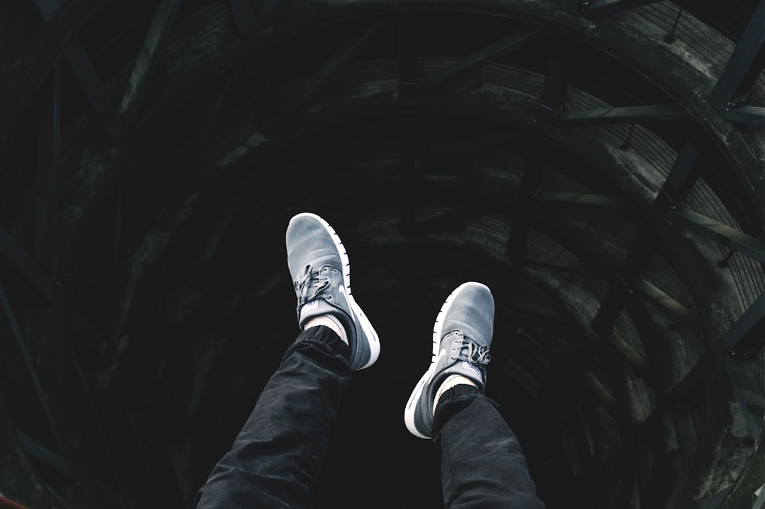 A person wearing white sneakers is standing on the edge of an abyss, with black jeans and gray shoes, looking down at their feet from above. The perspective includes low angle shots, dark tones, wideangle lenses, high contrast lighting, fast shutter speed, full body photos, loneliness.,,in –ar 128:85