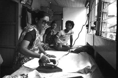 A black and white photo of an Indian woman ironing at a table in her small apartment, while two young men watch from behind her. They have sunglasses on and wear T-shirts and jeans. There is lots of sunlight and the walls are wooden. --ar 128:85