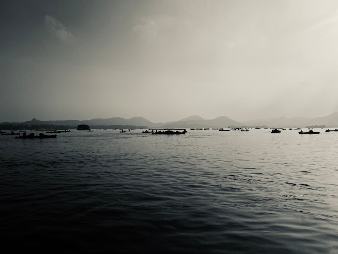 In the distance, there is an endless sea with boats floating on it. In front of them lies West Lake in Hangzhou and distant mountains. The sky above has dark clouds. There was no one around, only fishingmen were working at their fishermen’s seats. A black and white photo captures this serene scene. –ar 4:3