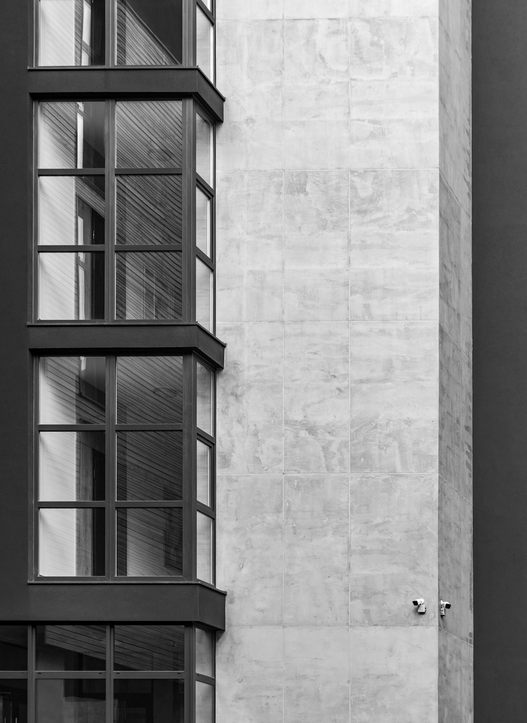 A black and white photo of the corner exterior wall of an office building with modern window frames, a tall concrete pillar in front of it. It is an architectural photography showing architectural detail, taken with a Leica M6 camera and macro lens at an aperture of f/2.0. Soft natural lighting and a minimalistic composition create an architectural mood, with a neutral color palette depicting a modern urban environment. –ar 93:128