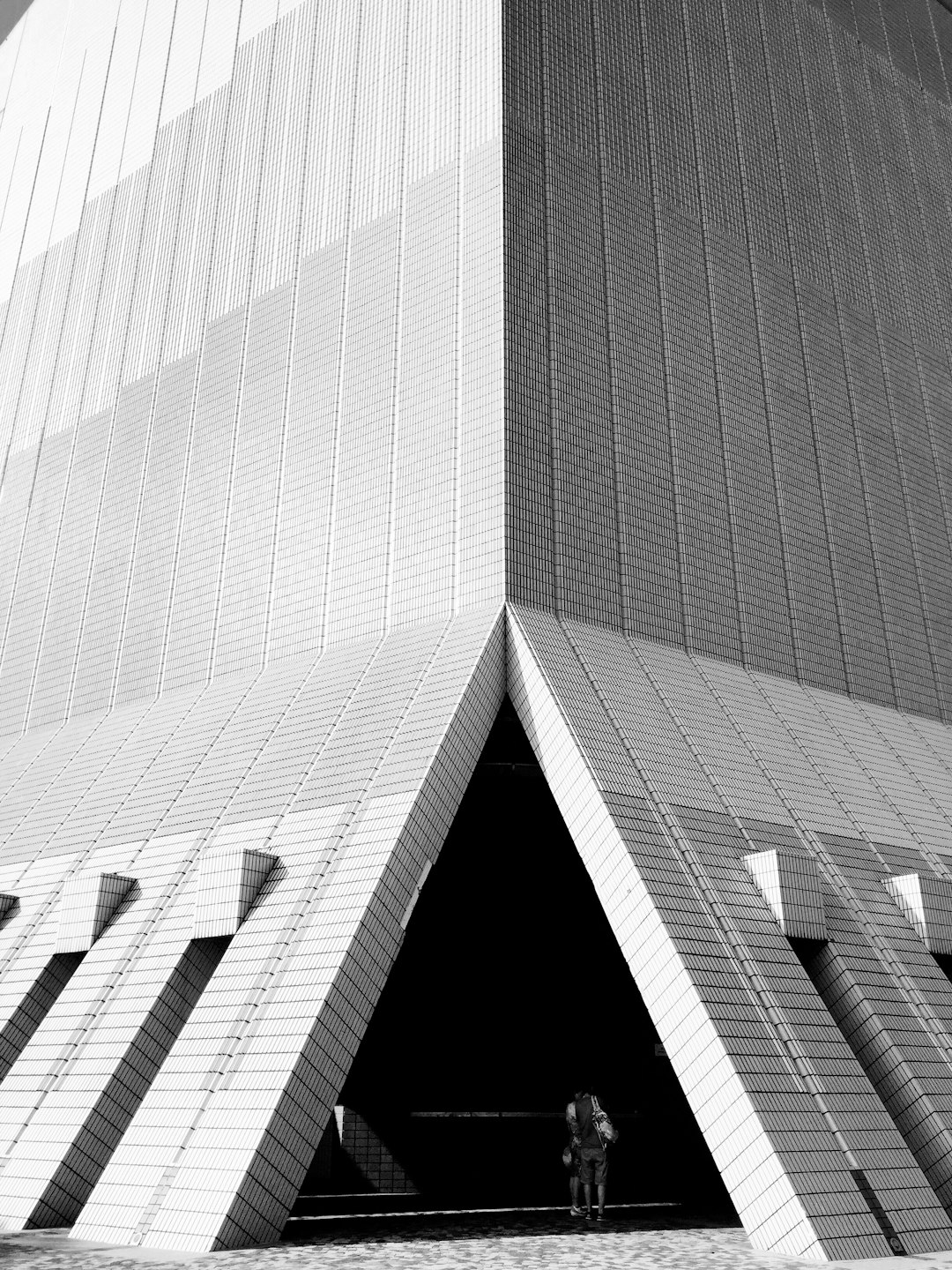 The entrance of the concert hall features triangular shapes, which can be seen in detail. The building is made up entirely from white bricks and has sharp edges on its sides. A couple stands at an angle near it. Black and white photography, low angle shot. –ar 3:4