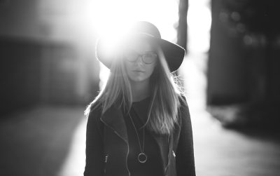 Black and white street photography of a young woman with long blonde hair, wearing glasses and a hat, standing on the sidewalk in downtown Los Angeles. She is looking at the camera. The sun shines through her, creating a lens flare. Shot in the style of arriflex 35 BL Camera, Canon K35 Prime Lenses, 70mm. --ar 8:5