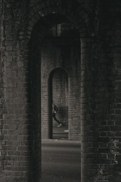 A black and white photograph shows an arched brick tunnel with two openings, taken from the perspective of someone running through it towards the camera. In one of the openings sits another person on a small bench in dark . The tunnel is dark and gloomy with high contrast. --ar 85:128