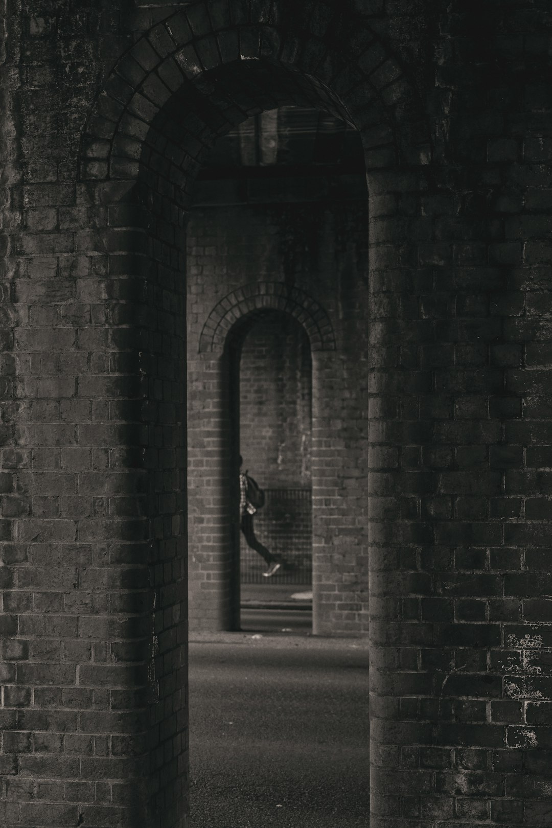 A black and white photograph shows an arched brick tunnel with two openings, taken from the perspective of someone running through it towards the camera. In one of the openings sits another person on a small bench in dark . The tunnel is dark and gloomy with high contrast. –ar 85:128