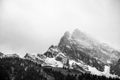 Black and white photography of the top part of The Grand Teton mountain range in winter, in the style of [Ansel Adams](https://goo.gl/search?artist%20Ansel%20Adams). There is snow on some parts of it. It's a cloudy day with fog covering much of the scene. Mountain peaks tower above dense forest below them. High contrast black and white with misty tones. Low angle shot looking up at the mountains. --ar 128:85