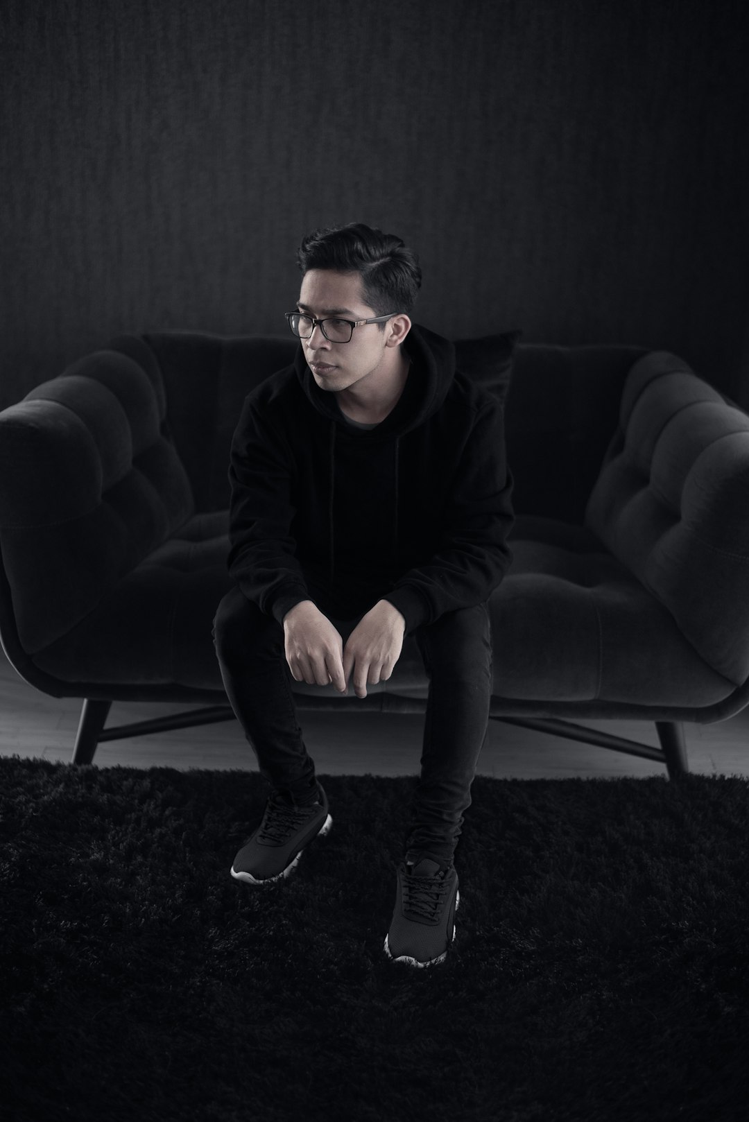 full body photography of an Asian man wearing a black hoodie and glasses, sitting on the couch with his legs crossed in front, against a dark background with a black carpet floor and black furniture. The photograph is in the style of by an unknown artist. –ar 85:128