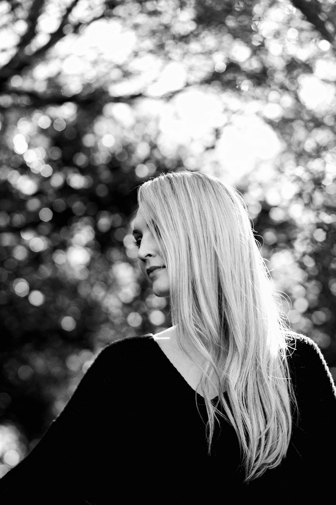 portrait of blonde woman in her thirties, black and white photography, blurry trees background, happy expression, side view, long hair, bokeh, high contrast, soft light, Hasselblad X2D 65mm lens –ar 85:128