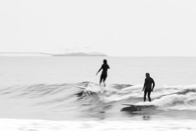 A black and white photo of surfers in the distance on waves, taken from the beach with a blurred foreground, using depth of field effects, in a minimalist style, with low contrast and saturation, captured with a Leica M6 camera, sparsely composed, with depth of field techniques, utilizing a long exposure, in the style of a double exposure, evoking summer vibes, featuring surfboards and a surfer. --ar 128:85