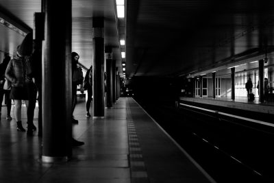 A black and white photo of people waiting at the station, with shadows on one side of an empty platform. The scene is captured in high contrast with sharp lines and soft lighting, creating a dramatic effect that emphasizes the solitude against the bustling atmosphere outside. --ar 128:85